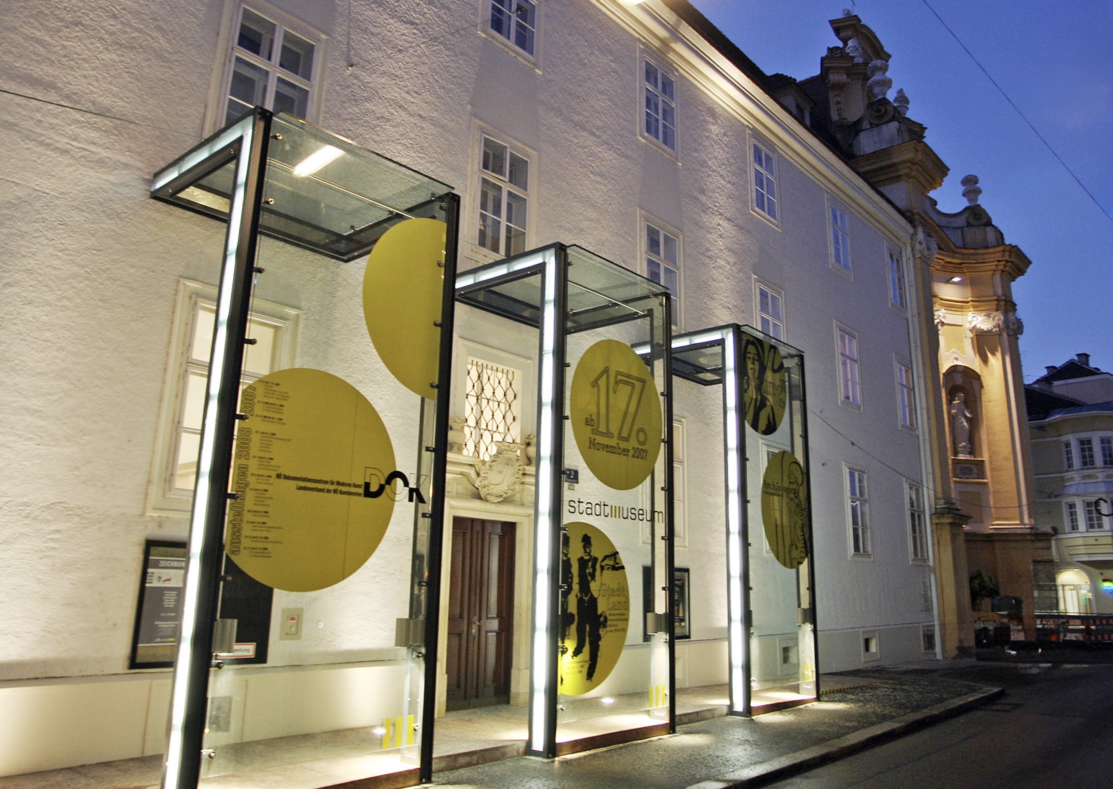 Außenansicht des Stadtmuseums St. Pölten bei Nacht mit beleuchteten Vitrinen und gelben Grafiken