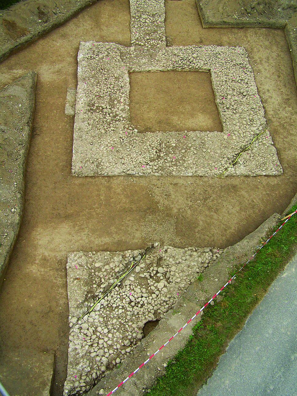 Luftaufnahme der Ausgrabung des Südtors des Legionslagers Albing: der rechteckige Grundriss des steinernen Torhauses mit Innenhof und vorgelagerter Straße ist freigelegt; ein rot‑weißes Vermessungsband markiert die Grabungsfläche, umliegender Kies und Erde bilden den Kontrast.