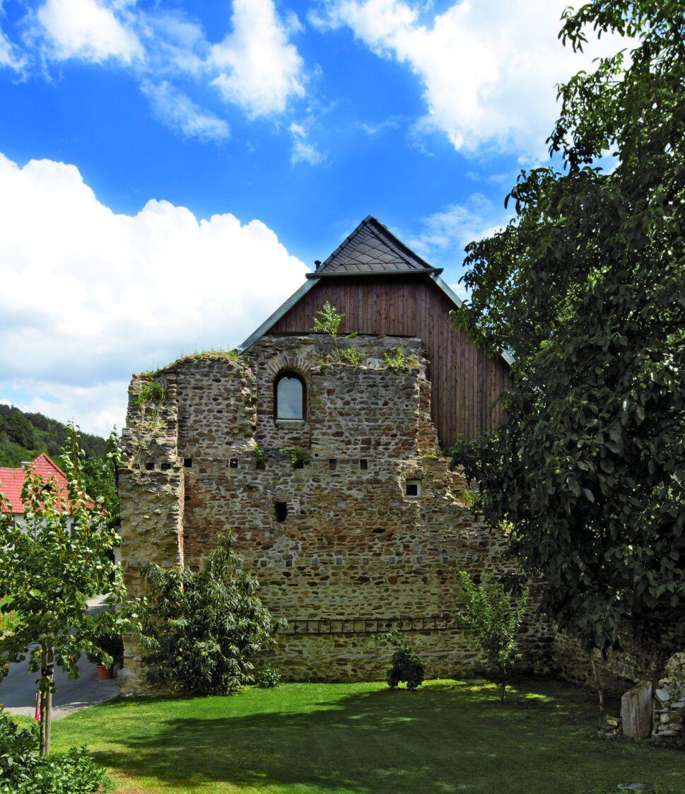 Ruine des römischen Wachturms von Bacharnsdorf: halbrundes, mehrgeschossiges Bauwerk aus unregeläßigem Bruchsteinmauerwerk mit kleinen Fensteröffnungen, teilweise überragt von einem modernen Satteldach. Im Vordergrund ein gepflegter Garten mit Gras, Baum und Büschern; dahinter erheben sich die Ruine und der bewaldete Hang an der Donau unter blauem Himmel mit Wolken.