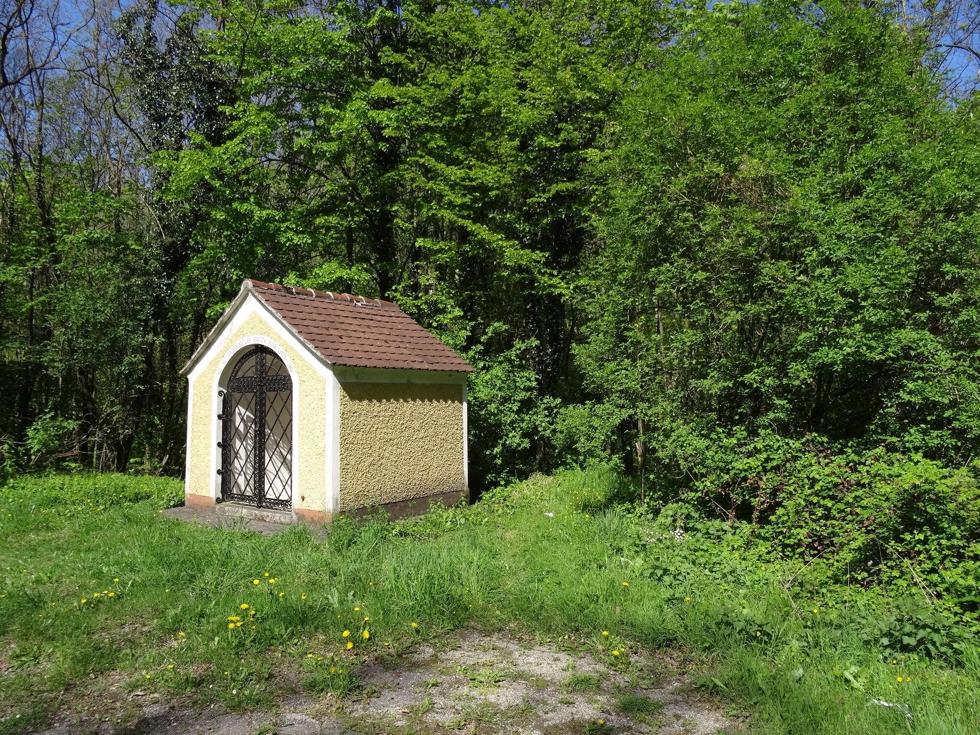 Fundstelle Blashaushgraben: kleines, gelb verputztes Kapellenhäuschen mit ziegelgedecktem Satteldach und schwarzem Gittertor markiert die Stelle eines römischen Wachturms; umgeben von dichten Laubbäumen, Gras und blühendem Löwenzahn.