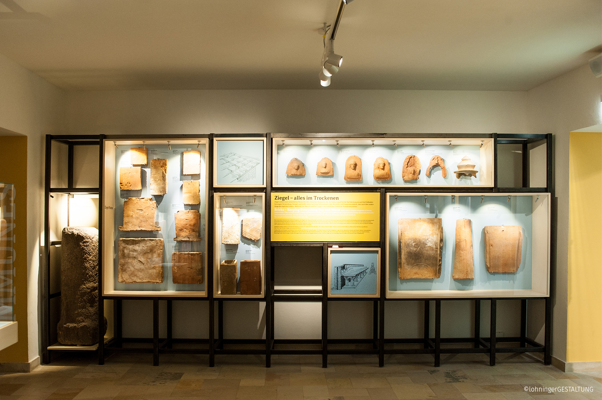 Ansicht der Dauerausstellung im Museum Lauriacum in Enns: eine große Vitrine mit dunklem Metallrahmen und beleuchteten Regalen präsentiert antike Ziegel, Inschriftensteine und Reliefplatten; in der Mitte ein gelb hinterlegter Informationstext, rechts und links Stelen und Exponate, alles in einem hell ausgeleuchteten Ausstellungsraum.