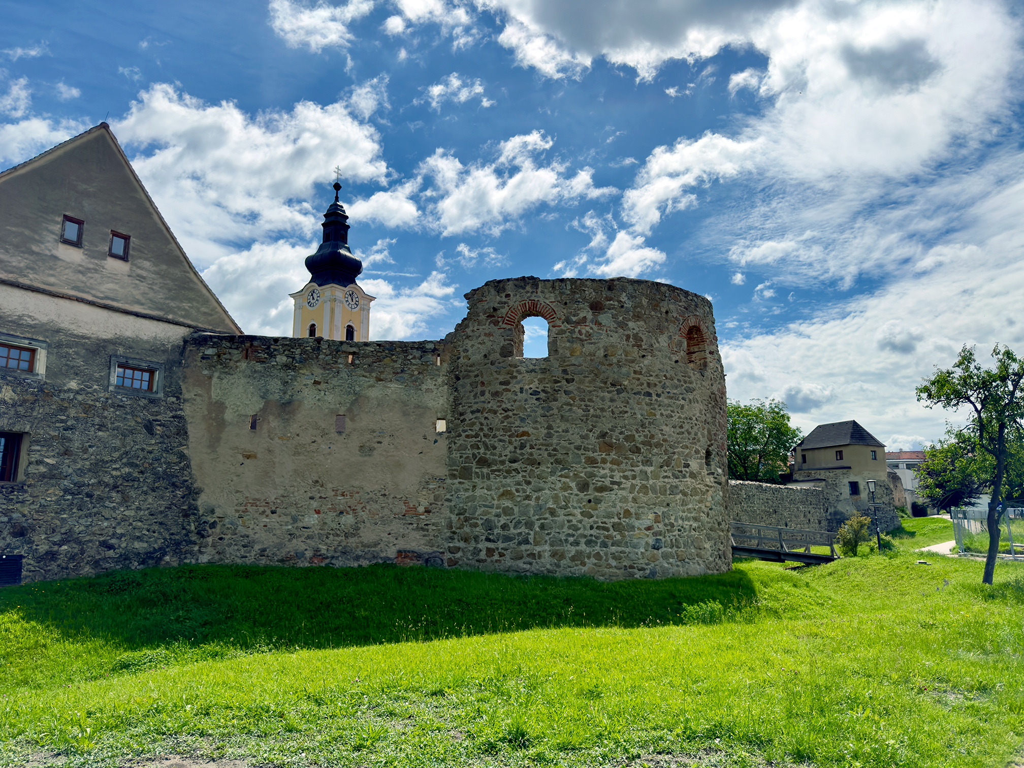 Der spätantike Hufeisenturm in Mautern an der Donau: massiver hufeisenförmiger Turm aus Bruchsteinmauerwerk mit kleinem Fenster, Teil der Kastellmauer; davor Rasenfläche, dahinter Pfarrkirche und moderne Häuser.