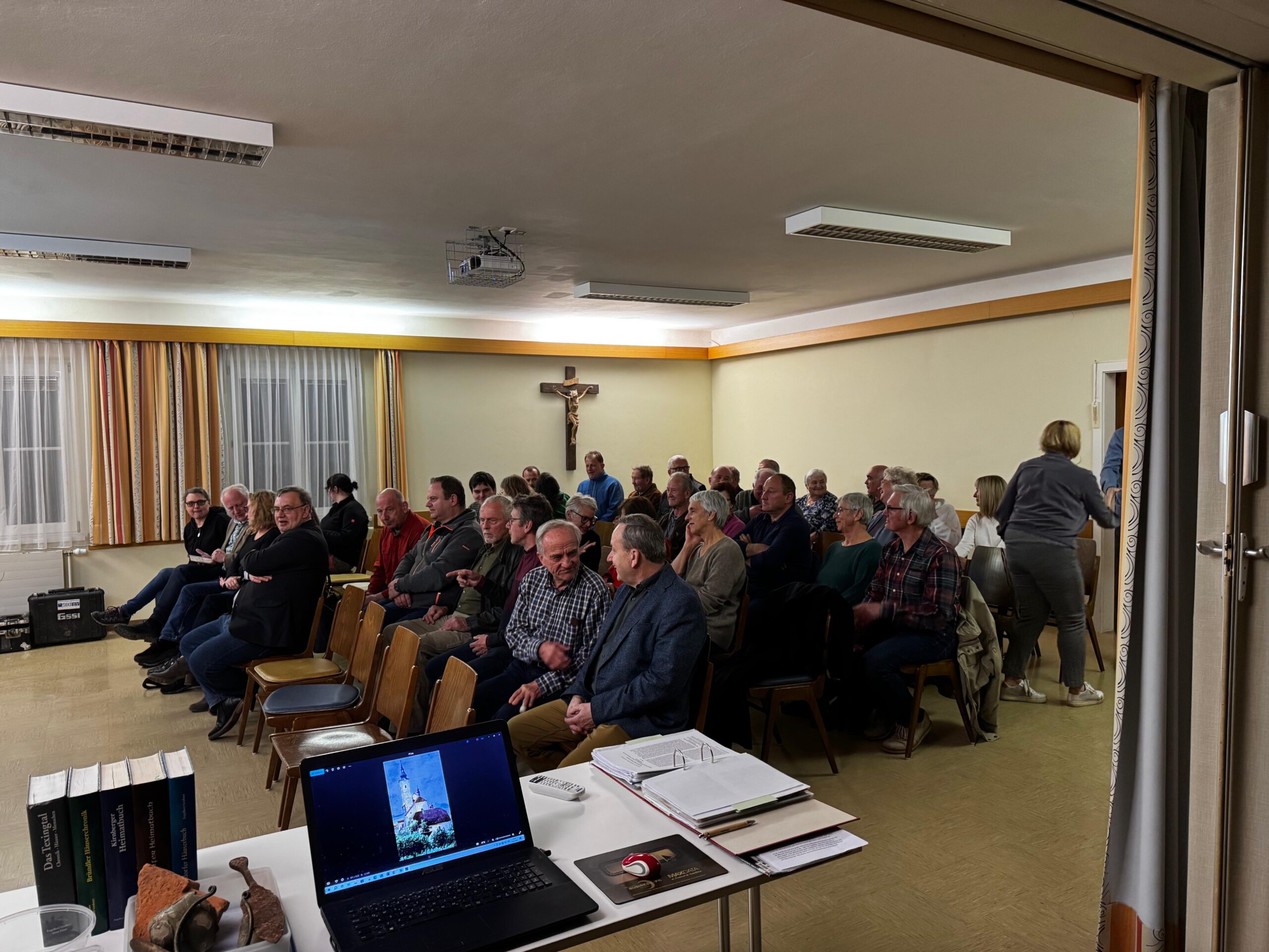 oberndorf-vortrag-2025 Publikum im Pfarrsaal von Oberndorf an der Melk hört 2025 einen Vortrag über römische Geschichte: Die Besuchenden sitzen in Reihen vor einem Tisch mit Laptop, Büchern und Keramikfragmenten, im Hintergrund gelbe Vorhänge und ein Kreuz an der Wand.