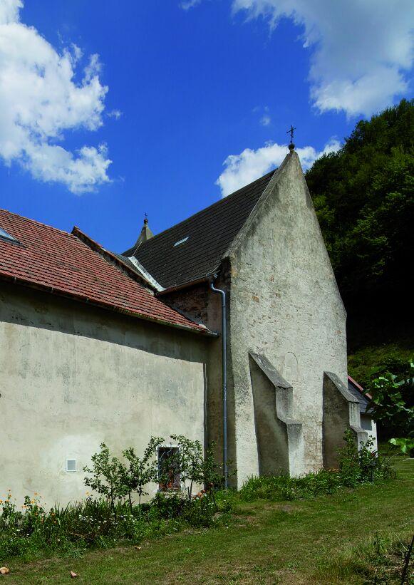 Kirche und angebautes Wohnhaus von Sankt Lorenz teilen sich die Mauern des spätantiken Wachturms: Weiß verputzte, hoch aufragende Wände mit Strebepfeilern, rotem Ziegeldach und angrenzendem Hang.