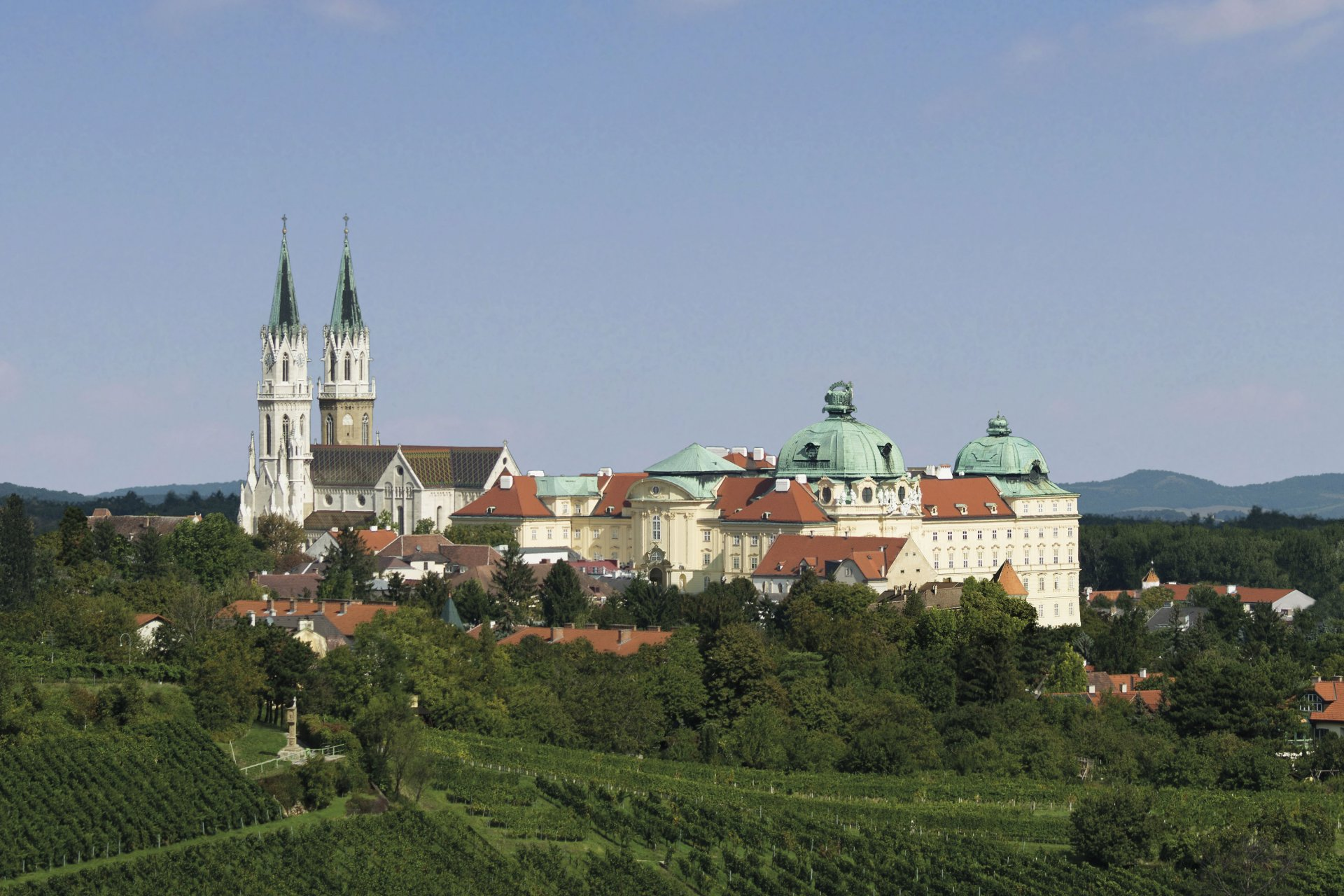 Panoramablick auf das Stift Klosterneuburg bei Wien: die barocken Stiftsgebäude mit grünen Kuppeln und die gotische Kirche mit zwei hohen Türmen erheben sich über terrassierten Weingärten; im Vordergrund Weinreben und Bäume, im Hintergrund der Wienerwald unter blauem Himmel.