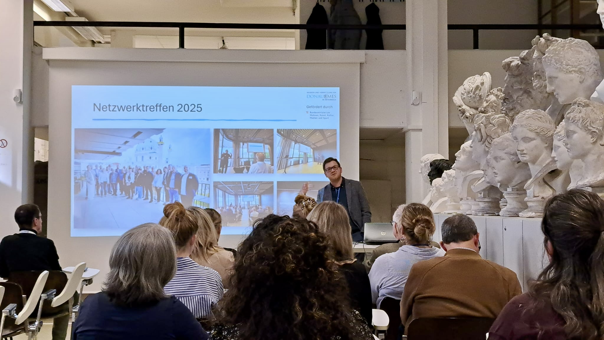 Vortrag von Dominik Hagmann beim 20. Österreichischen Archäologietag 2026 am Institut für Klassische Archäologie der Universität Wien. Er steht vor einer projizierten Präsentationsfolie mit dem Titel „Netzwerktreffen 2025“ und Bildern vergangener Veranstaltungen und spricht zu einem sitzenden Publikum in einem Hörsaal- bzw. Institutsraum. Rechts im Bild befinden sich mehrere dicht aufgestellte weiße Gipsabgüsse antiker Porträtbüsten. Der Vortrag behandelt die Arbeit des Vereins „Museen und Vermittlung am Donaaulimes“. Foto: Nisa Iduna Kirchengast
